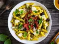 Penne con pesto, pomodori secchi, olive nere, capperi e scaglie di parmigiano. Un piatto colorato e saporito.