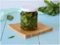 Un barattolo di olio al basilico fatto in casa, con foglie fresche immerse nell'olio dorato, su un tagliere di legno.