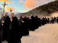 Monks in black robes process with a cross and lanterns, their solemn religious chants echoing as they walk towards a building at sunset.