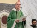 A man with a Benedictine title, wearing a green chasuble, speaks into a microphone while holding a paper.