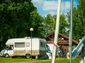 Słoneczny dzień na kempingu na Mazurach. Kamper stoi na trawie obok drewnianego budynku, w tle zielone drzewa i plac zabaw.