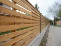 Ett modernt, liggande staket i trä med metallstolpar. Staketet löper längs en trottoar och en grönskande gräsmatta.