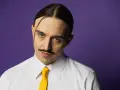 Tommy Cash w białej koszuli i żółtym krawacie, z charakterystycznym wąsem i przedziałkiem na środku głowy, na fioletowym tle.