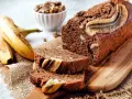 Saftiges, veganes Bananenbrot mit Walnüssen und Bananenscheiben, daneben frische Bananen und eine Schüssel Nüsse.