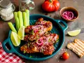 Skrzydełka z BBQ w sosie, podane z łódeczkami limonki, selerem i marynowaną cebulką. Obok grzanka i pomidorki.