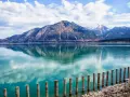 Spiegelglatter See mit Bergen im Hintergrund, wie man ihn in Bayern sehen kann. Holzpfähle ragen aus dem türkisfarbenen Wasser.