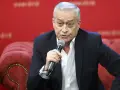 Janusz Szydłowski przemawia do mikrofonu, siedząc na czerwonej, pikowanej kanapie.