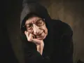 Starszy mężczyzna w czarnym kapturze, z okularami na nosie, opiera głowę na dłoni. Jego twarz wyraża głębokie cierpienie i zadumę.