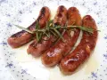 Salchichas al vino, doradas y jugosas, reposan sobre un plato blanco con delicados motivos azules, adornadas con ramitas de romero fresco.