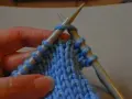En hand håller stickor med blått garn. En rätmaska formas på stickan, redo att stickas.