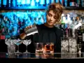 Barman nalewa drink z shakera do szklanki, w tle półki z butelkami i niebieskie światła.