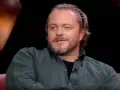 Tomasz Schuchardt, znany z wielu filmów, seriali i programów, z charakterystyczną fryzurą i niebieskimi oczami, siedzi na kanapie.
