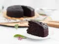 Ein saftiges Stück veganer Schokokuchen liegt auf einem Teller. Im Hintergrund ein ganzer Kuchen, dekoriert mit grünen Blättern und roten Beeren.