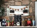 Stół z napojami i składnikami do drinków, tworzący domowy co zamiast drink baru. Na półce stoją butelki alkoholu, syropy, kartony z sokami i dwa duże słodkie poncze.