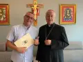 Uśmiechnięty mężczyzna z dokumentem i biskup pozują na tle krzyża i obrazów. Mężczyzna trzyma dokument z herbem, a biskup ma na sobie sutannę.