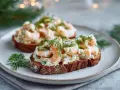 Två toast Skagen med dill på mörkt bröd, serverade på en tallrik med juldekorationer i bakgrunden.