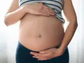 Brzuszek w ciąży z widocznym pępkiem, otoczony dłońmi przyszłej mamy.