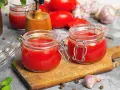 Deux bocaux en verre remplis de coulis de tomates, sur une planche en bois. Des tomates, de l'ail et du basilic complètent la scène.