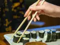 Dłoń z pałeczkami sięga po rolkę sushi. Na talerzu ułożone są kawałki sushi, każdy z nich to mała porcja smaku i kalorii.