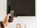 Dłoń trzymająca pilot do telewizora na tle wyłączonego ekranu.