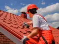 Pracownik w czerwonym kasku i ogrodniczkach dokonuje wymiany dachu, przykręcając nowe dachówki wkrętarką.