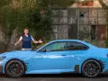 Mężczyzna prezentuje błękitne BMW M2 G87 przed zniszczonym budynkiem.