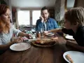 Rodzina przy stole, pizza, w tle mężczyzna zamyślony. Czasem nawet przy wspólnym posiłku pojawiają się kłótnie o dzieci z poprzedniego związku.