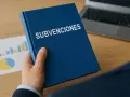 Mano sostiene un libro azul con "SUBVENCIONES" escrito. Detrás, gráficos y un portátil.