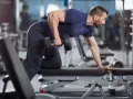 Man utför enarms hantelrodd på ett gym. Han stöttar sig mot en bänk med ena handen och drar en hantel mot bröstet med den andra.