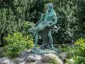 Bronzestatue eines Mannes, der eine Frau trägt, vor einem grünen Hintergrund mit einem kleinen Wasserfall in Berlin.