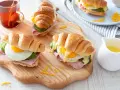 Trois croissants au jambon, œuf au plat, avocat et fromage râpé, servis sur une planche en bois avec une tasse de thé.