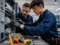 Giovane apprendista impara i segreti dei **corsi per elettricisti** con un esperto, lavorando su un quadro elettrico complesso.