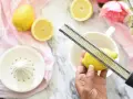 Hand reibt zeste einer zitrone über eine Reibe. Zitronen und Blumen liegen auf einem Marmortisch.