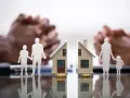 Figurines de famille séparées par une maison coupée en deux, symbolisant une convention d'indivision divorce. Mains floues en arrière-plan.