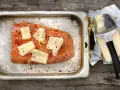 Lax i ugn, kryddad med chili och toppad med ostskivor, redo att tillagas. Bredvid ligger smör och en osthyvel.