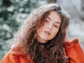 Jeune femme aux cheveux bouclés, portant une veste orange. La retouche photo netteté en ligne sublime son regard intense.