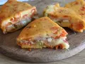 Fette di torta salata senza uova, ricca di pomodoro, formaggio filante e prosciutto, su un tagliere di legno.