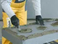 Pracownik w żółtym ubraniu i czarnych rękawicach rozprowadza zaprawę na styropianie, przygotowując go do klejenia.
