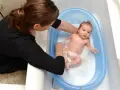 Mama delikatnie myje noworodka w niebieskiej wanience. Maluch pluska się w pianie, a mama uśmiecha się do niego.