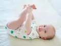 Ein süßes Baby, ca. 6 Monate alt, liegt auf dem Rücken und spielt mit seinen Füßen. Es trägt einen weißen Body mit grünen Fröschen.