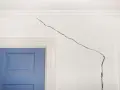Grosse fissure sur mur intérieur blanc, près d'une porte bleue. Il faut réparer cette fissure pour un intérieur impeccable.