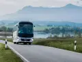 Biały **bus** marki **MAN** jedzie krętą drogą nad jeziorem, w tle góry.