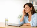 Donna con gli occhi che battono, appoggiata a un tavolo con un bicchiere d'acqua e limone e un laptop chiuso.