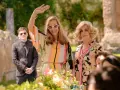 Due donne eleganti, una saluta con la mano, in un giardino. Un uomo in giacca scura fa da guardia. Un semplice favore per un evento speciale.