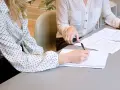 Two women in polka-dot shirts review documents. One takes notes, implying they are discussing who can see committee meeting minutes.