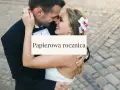 Para świętuje papierową rocznicę ślubu. Uśmiechnięci, w objęciach, z kwiatem we włosach panny młodej.