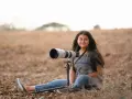 Młoda kobieta z długimi włosami siedzi na ziemi z profesjonalnym aparatem na statywie. Kompozycja otwarta podkreśla jej pasję do fotografii przyrodniczej.