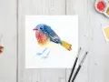 Un charmant dessin oiseau aquarelle aux couleurs vives, posé sur du papier blanc. Des feuilles d'automne et du matériel de peinture complètent la scène.