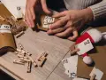 Händer som pysslar med en adventskalender. Små bruna paket med siffror och snöre, samt klädnypor och en tomteluva.