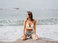 Uśmiechnięta kobieta w letniej stylizacji klęczy na plaży. Woda obmywa jej stopy, a fale tworzą piękne tło. To idealne, ładne pozy do zdjęć.